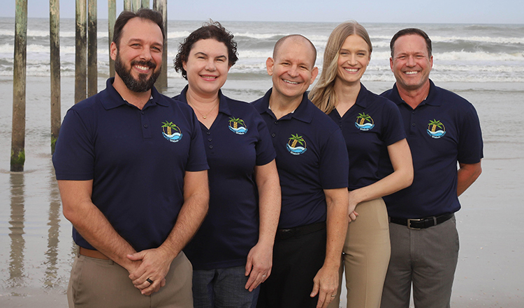 Five staff in blue Atlantic Wellness attire standing on the beach with waves in the background.